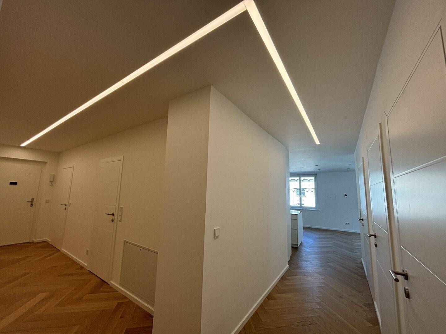 Langer, heller Flur mit modernem Lichtband an der Decke und Fischgrätparkettboden.