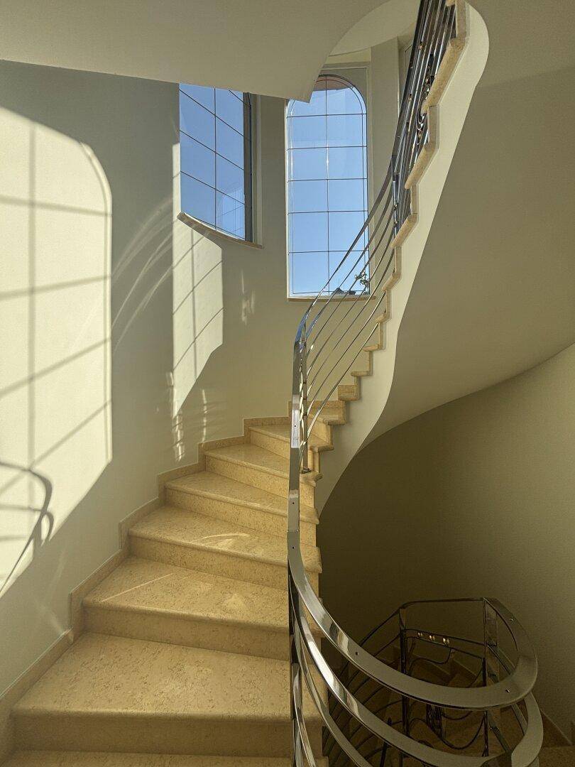 Elegante Wendeltreppe mit Marmorstufen und Metallgeländer, hell erleuchtet durch große Fenster.
