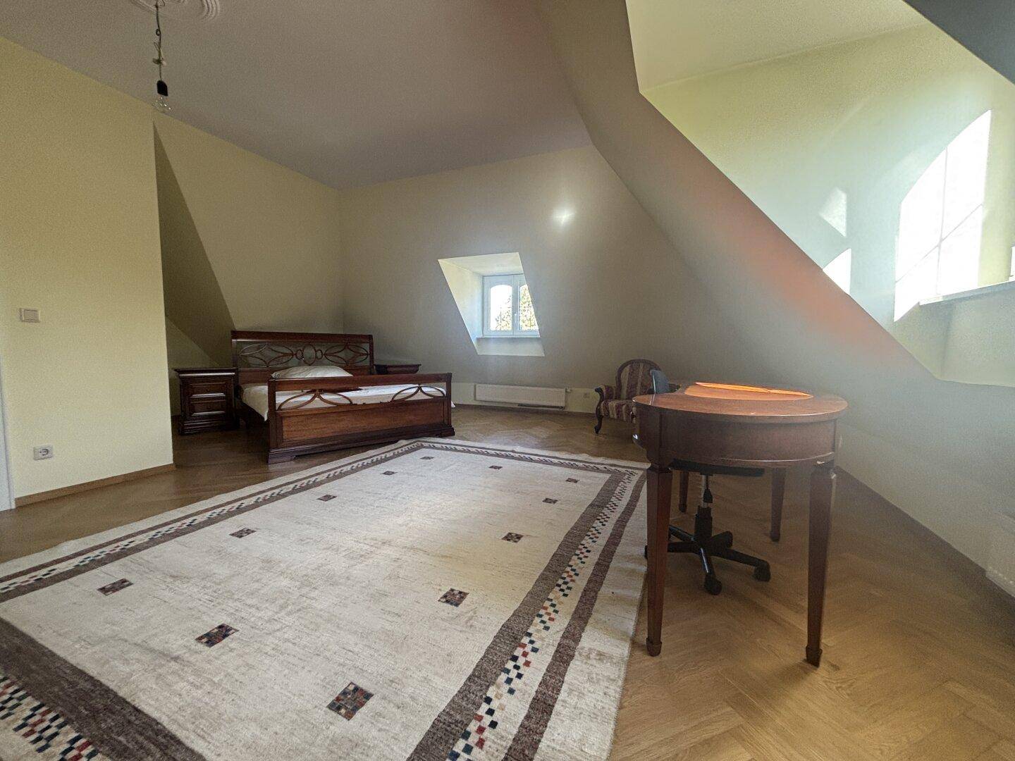 Geräumiges Schlafzimmer im Dachgeschoss mit Doppelbett, Schreibtisch und großem Teppich auf Parkettboden.
