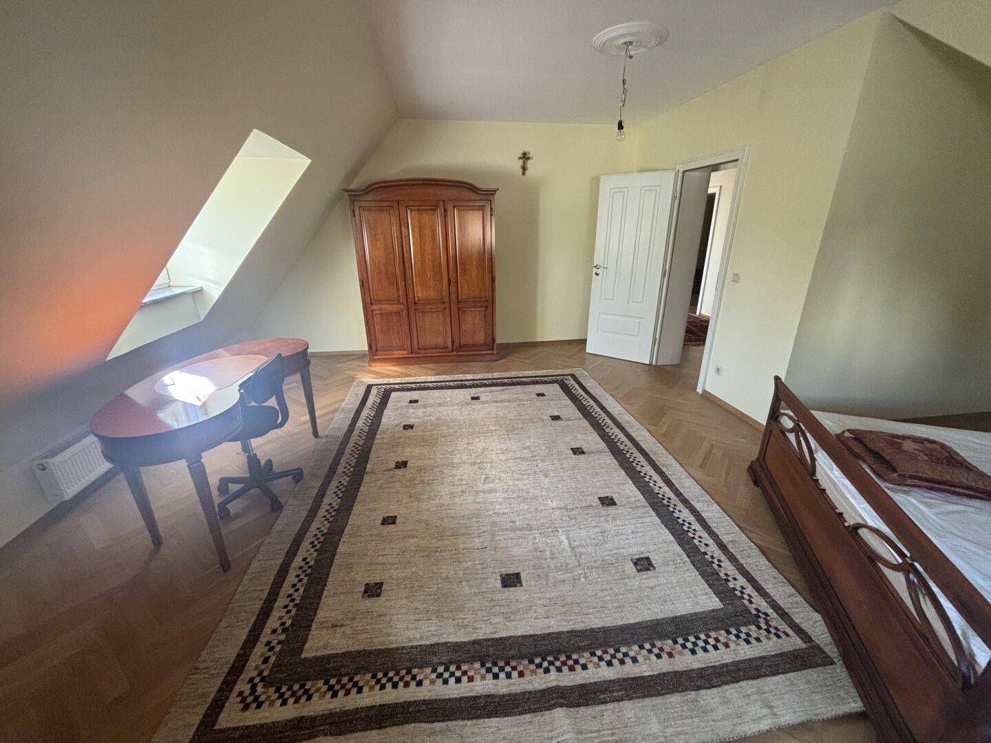 Großes Schlafzimmer im Dachgeschoss mit Holzschrank, Schreibtisch und Teppich auf Fischgrätparkett.