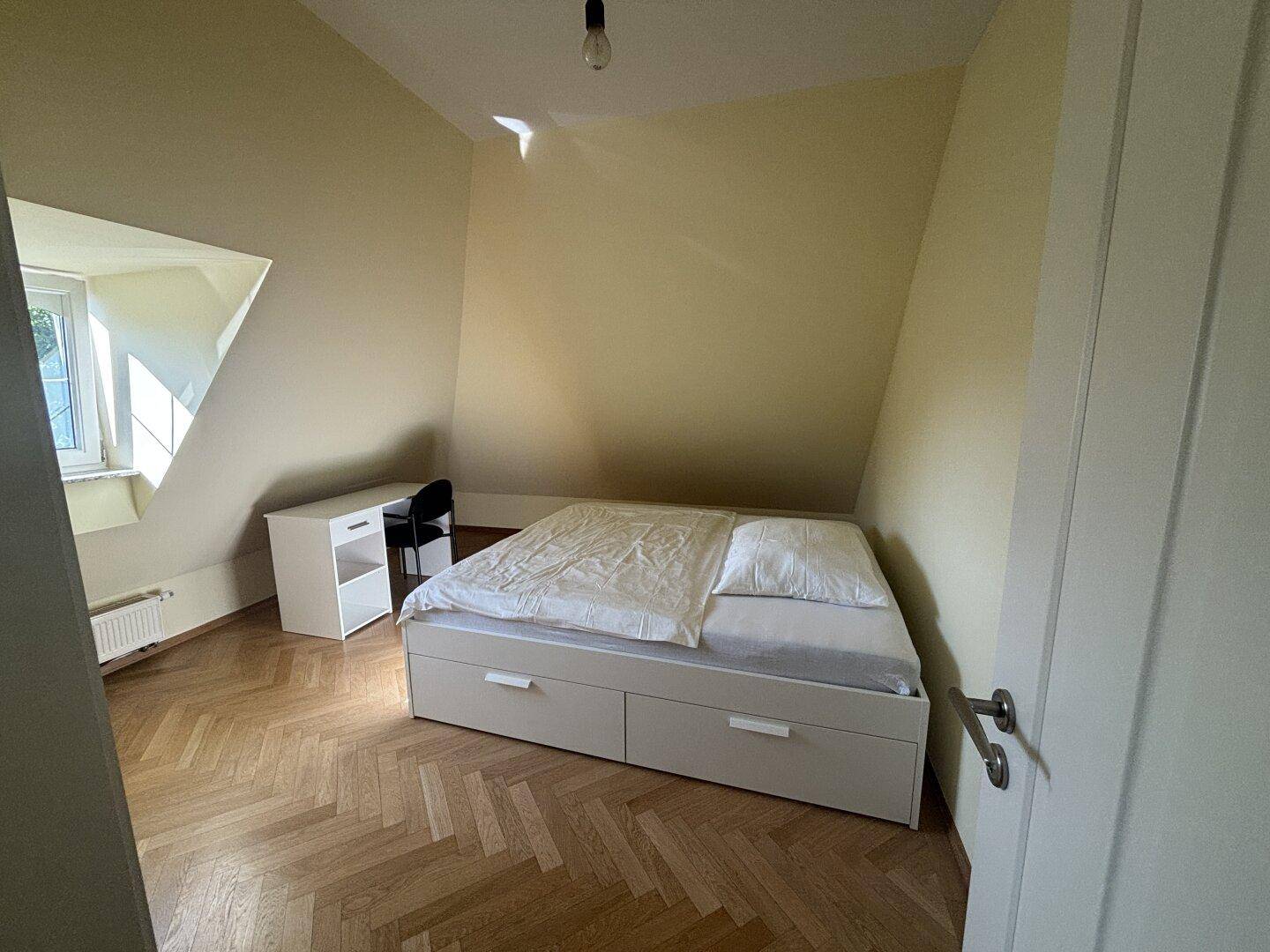 Gemütliches Schlafzimmer im Dachgeschoss mit Bett, Schreibtisch und Fischgrätparkett unter schrägen Decken.