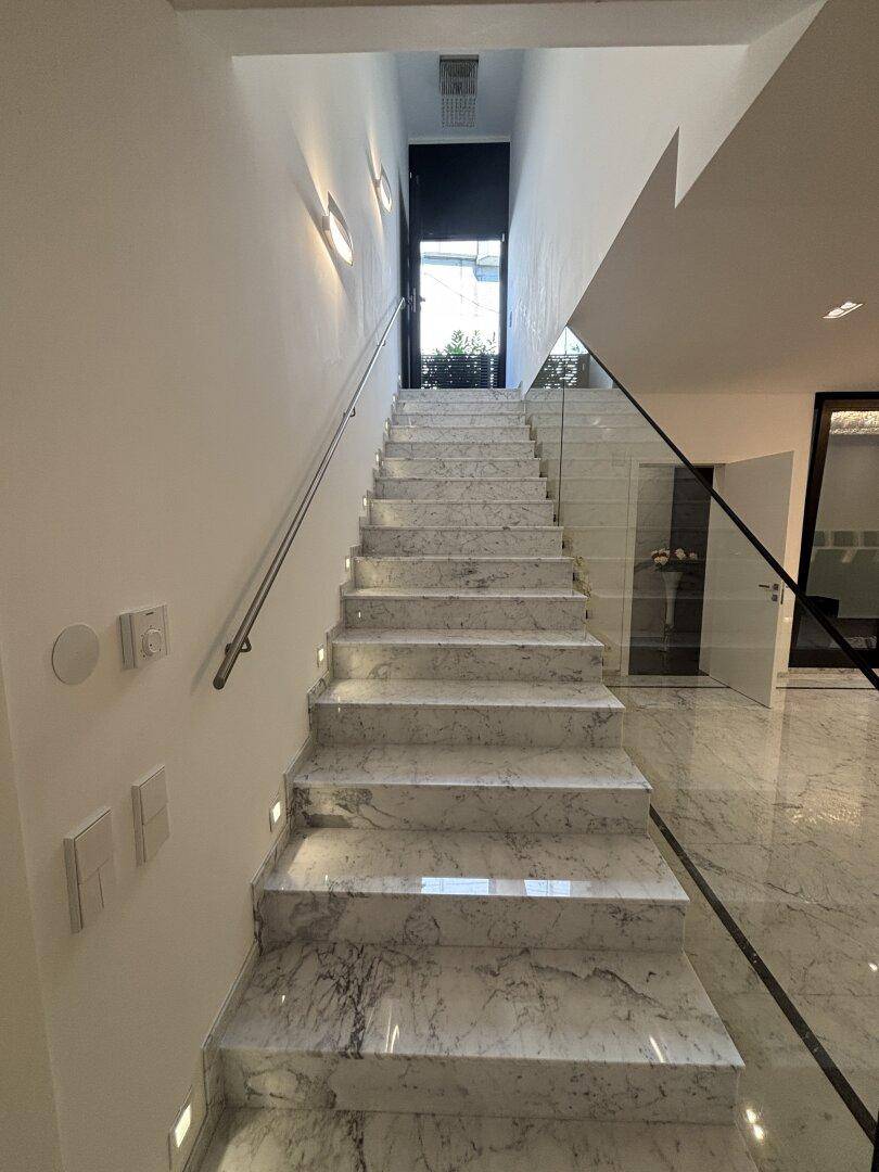 Moderne Treppe mit Marmorstufen, Glasgeländer und integrierter Beleuchtung, die zum Eingang führt.