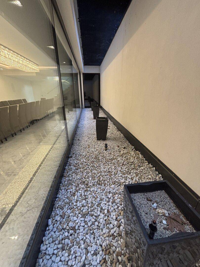 Schmaler Außenbereich mit Kieselsteinen und Pflanzenkübeln, angrenzend an eine große Glasfassade.