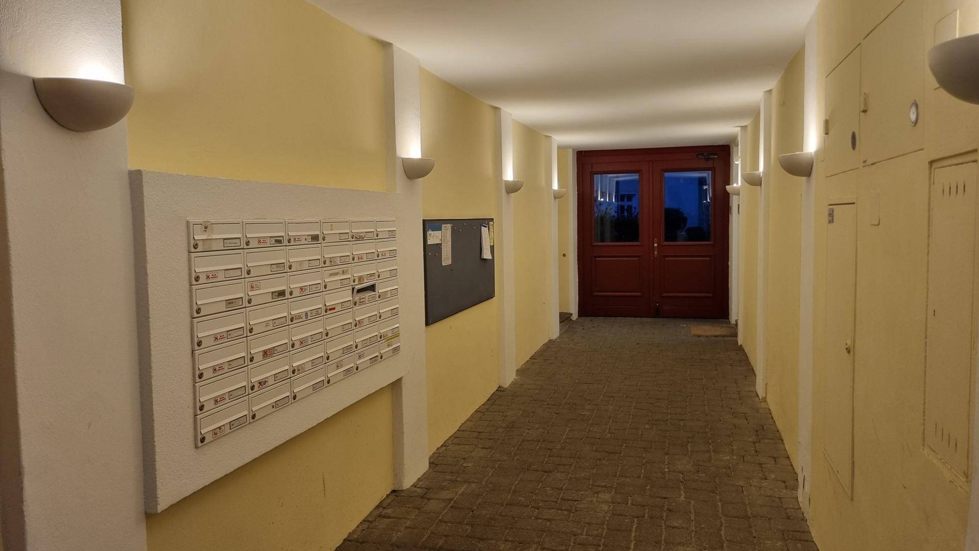 Heller Flur mit Briefkästen, gelben Wänden und einer roten Eingangstür am Ende.