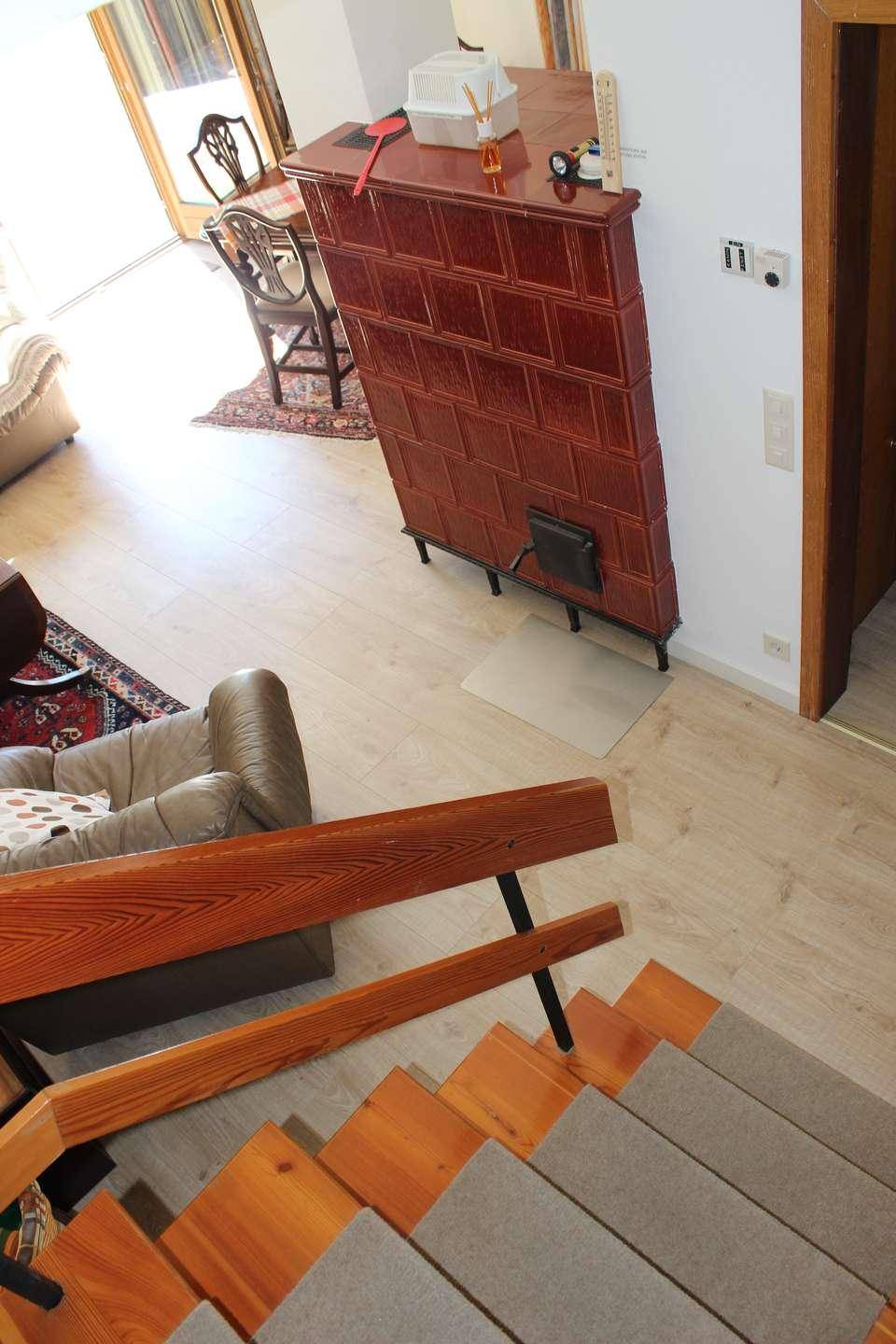 Treppenaufgang mit Holzgeländer und Blick auf den traditionellen Kachelofen im Wohnbereich.