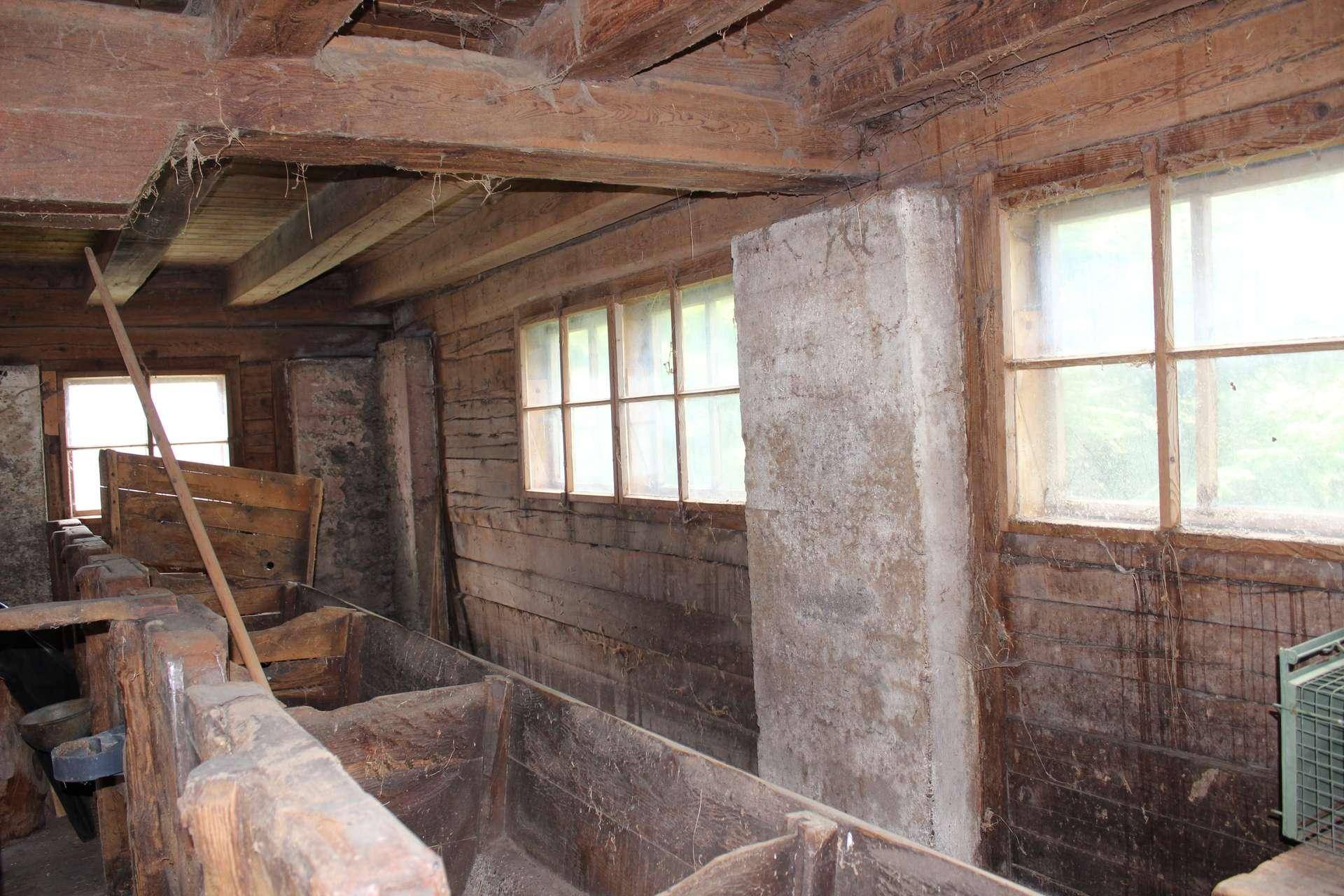 Innenansicht eines alten, rustikalen Stalls oder Scheune mit Holzwänden, kleinen Fenstern und Betonpfeilern.