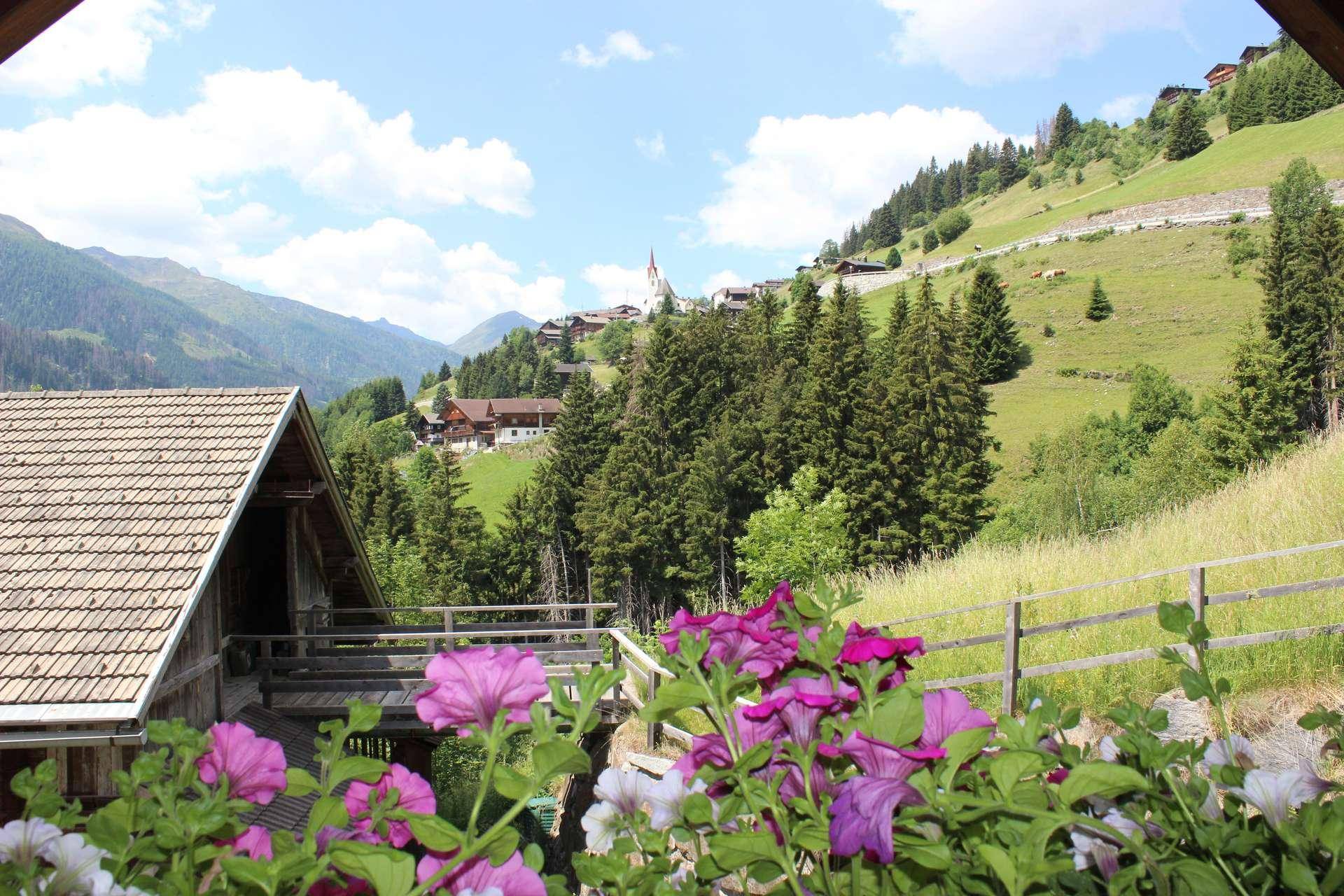 Blick von einem Balkon mit bunten Blumen auf ein malerisches Bergdorf und grüne Hügel.