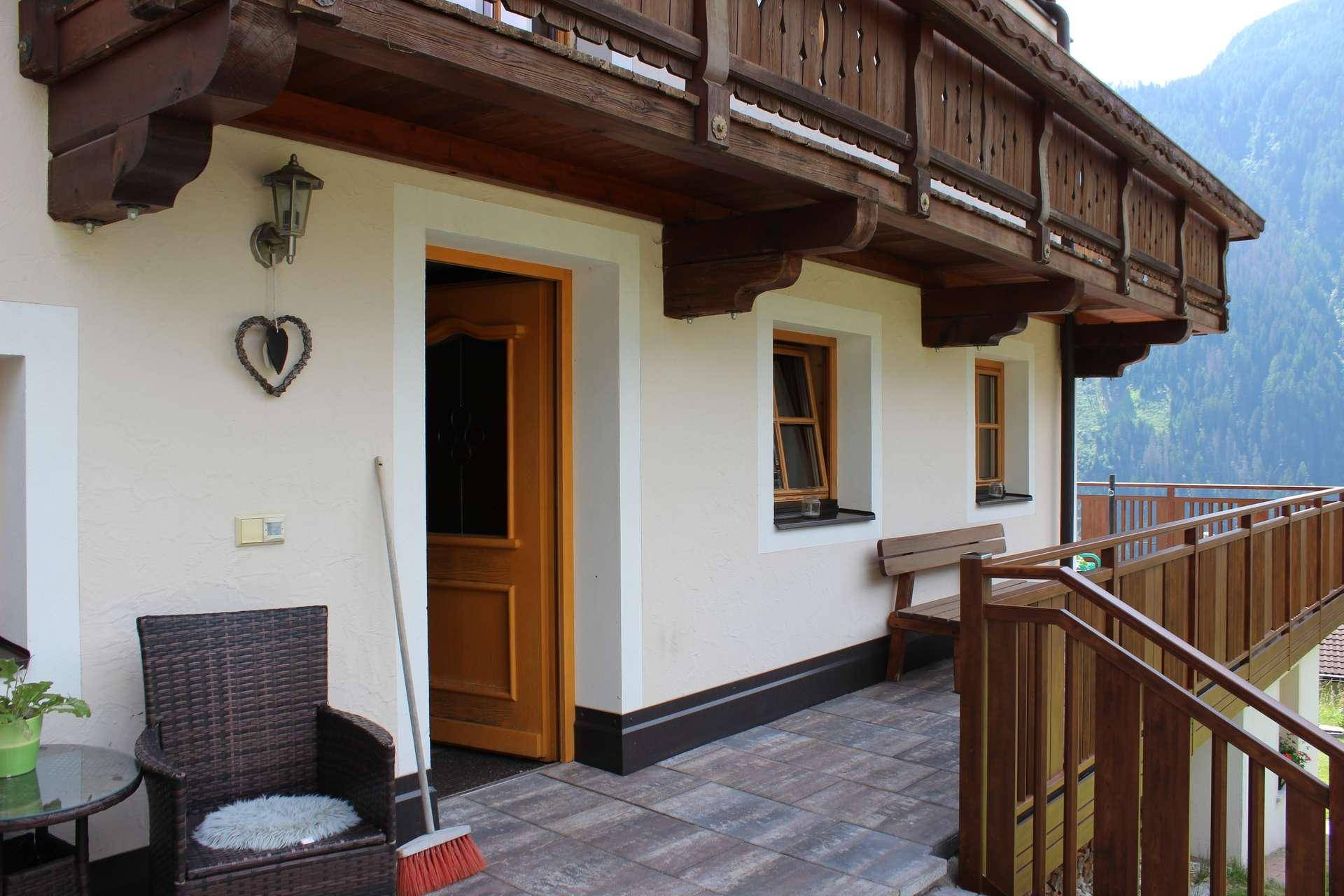 Außenbereich mit gepflasterter Terrasse, kleiner Sitzgruppe und Holzbalkon darüber, neben dem Hauseingang.