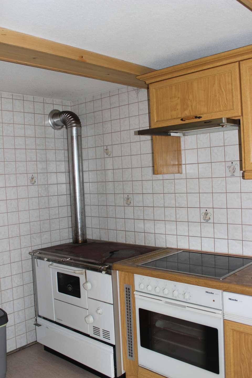 Küchenbereich mit traditionellem Holzofen, modernem Induktionskochfeld und Backofen, unter Holzbalken.