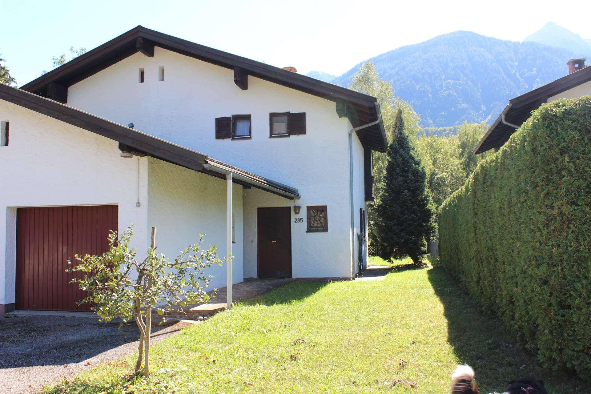 Vorderansicht des Hauses mit Garage, Vorgarten und Hecke, umgeben von Bergen.
