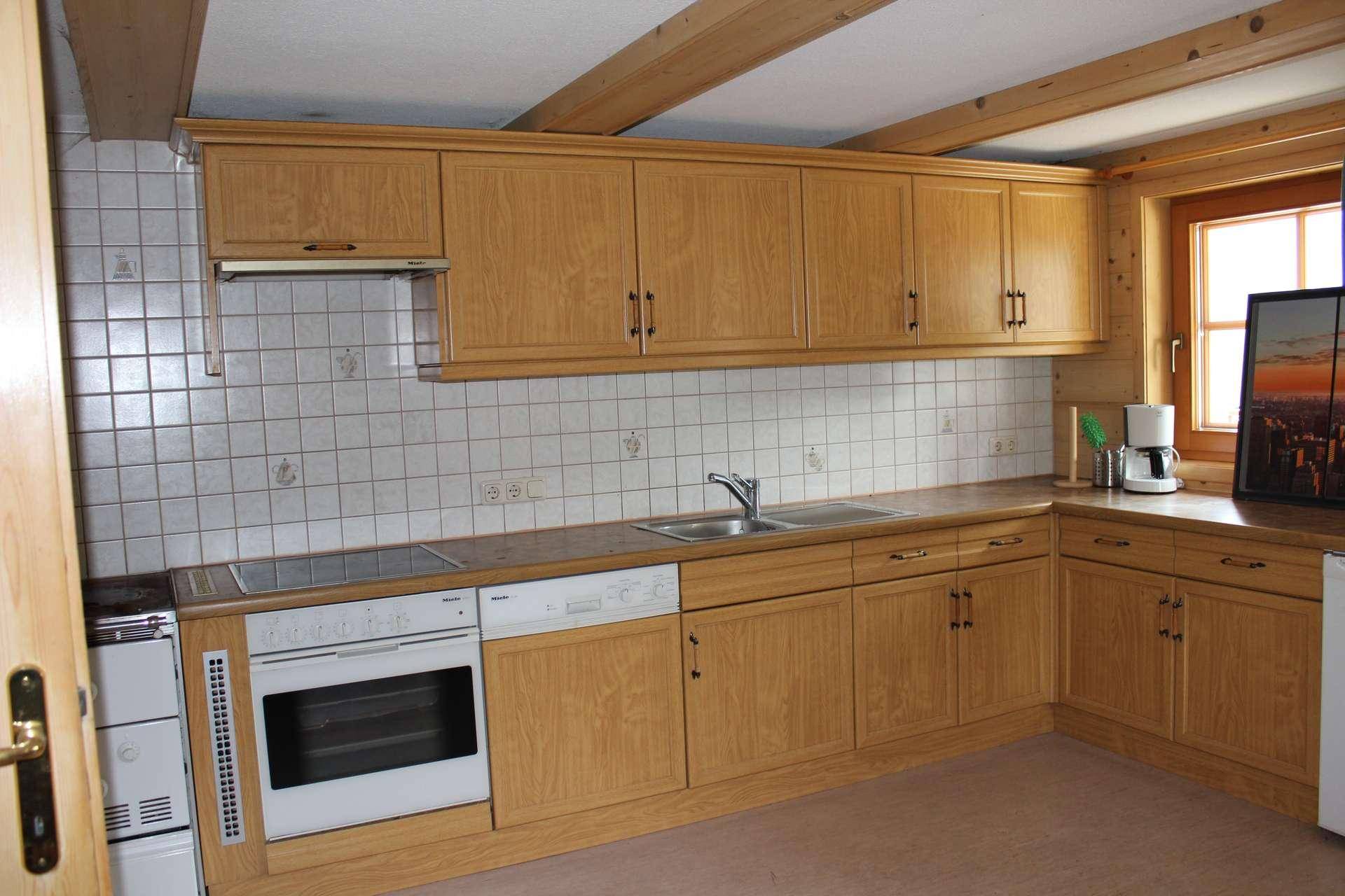 Küche mit hellen Holzschränken, weißem Fliesenspiegel, Einbauofen und Fenster, mit Holzbalken an der Decke.