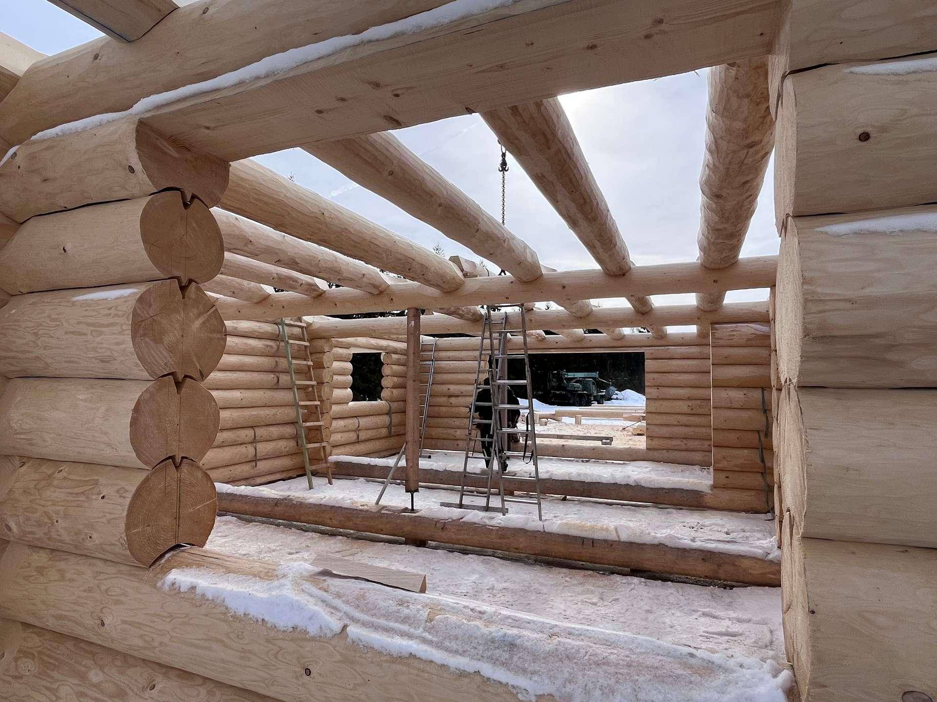 Innenansicht des im Bau befindlichen Blockhauses mit sichtbaren Holzstämmen und Deckenbalken.