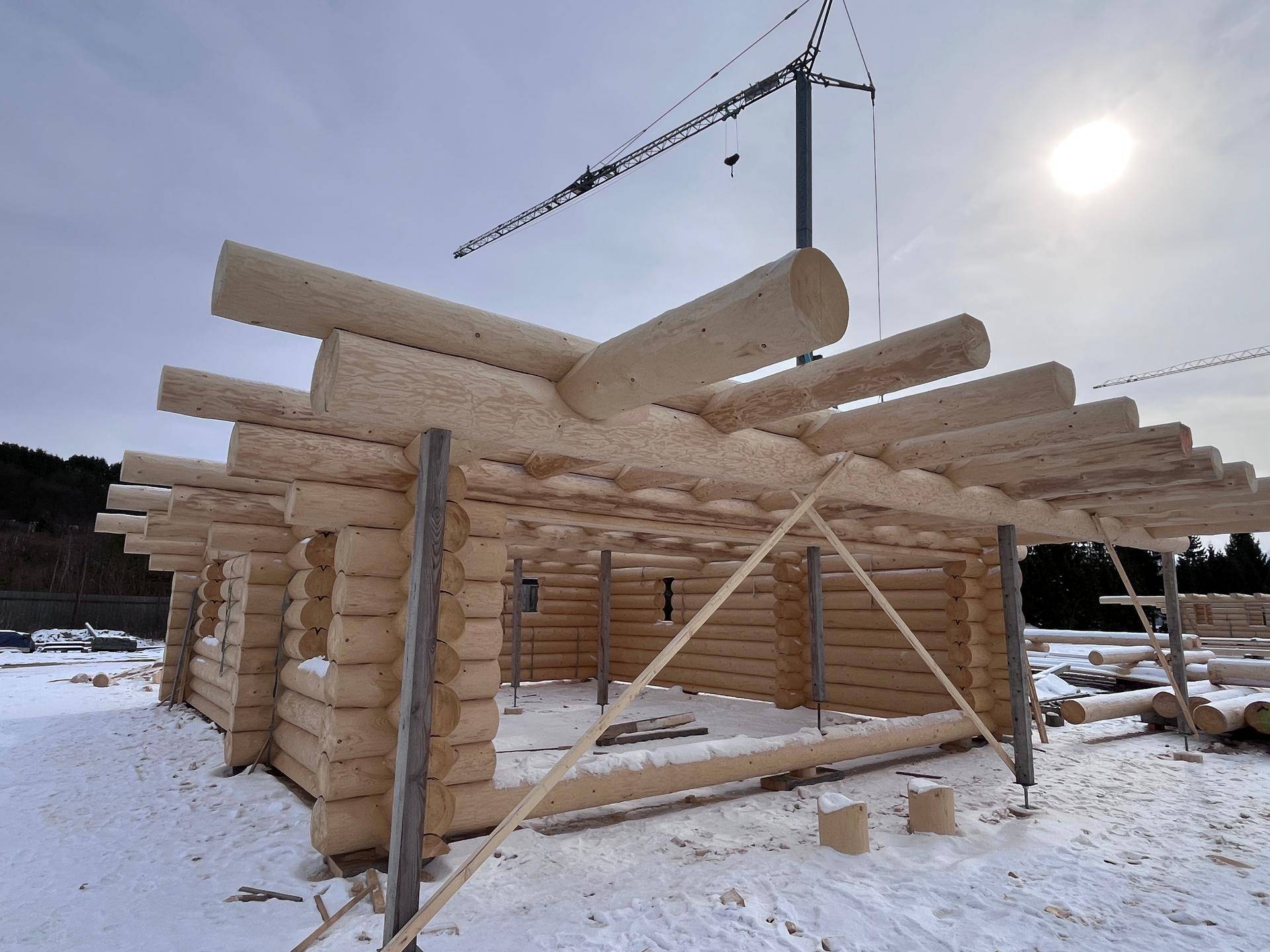 Baustelle eines Blockhauses im Winter mit sichtbaren Holzstämmen und Kran im Hintergrund.
