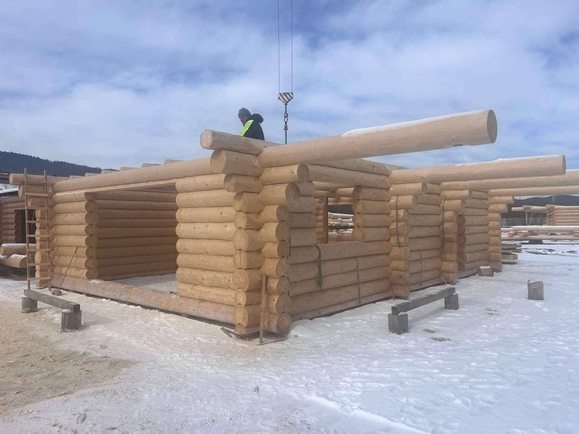 Baustelle eines Blockhauses im Winter, mit sichtbaren Holzstämmen und Bauarbeitern.