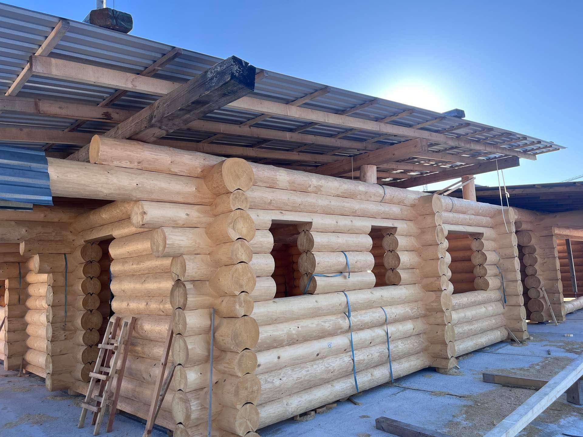 Fortschritt des Blockhausbaus mit gestapelten Holzstämmen und vorbereiteten Fensteröffnungen, bei Sonnenschein.