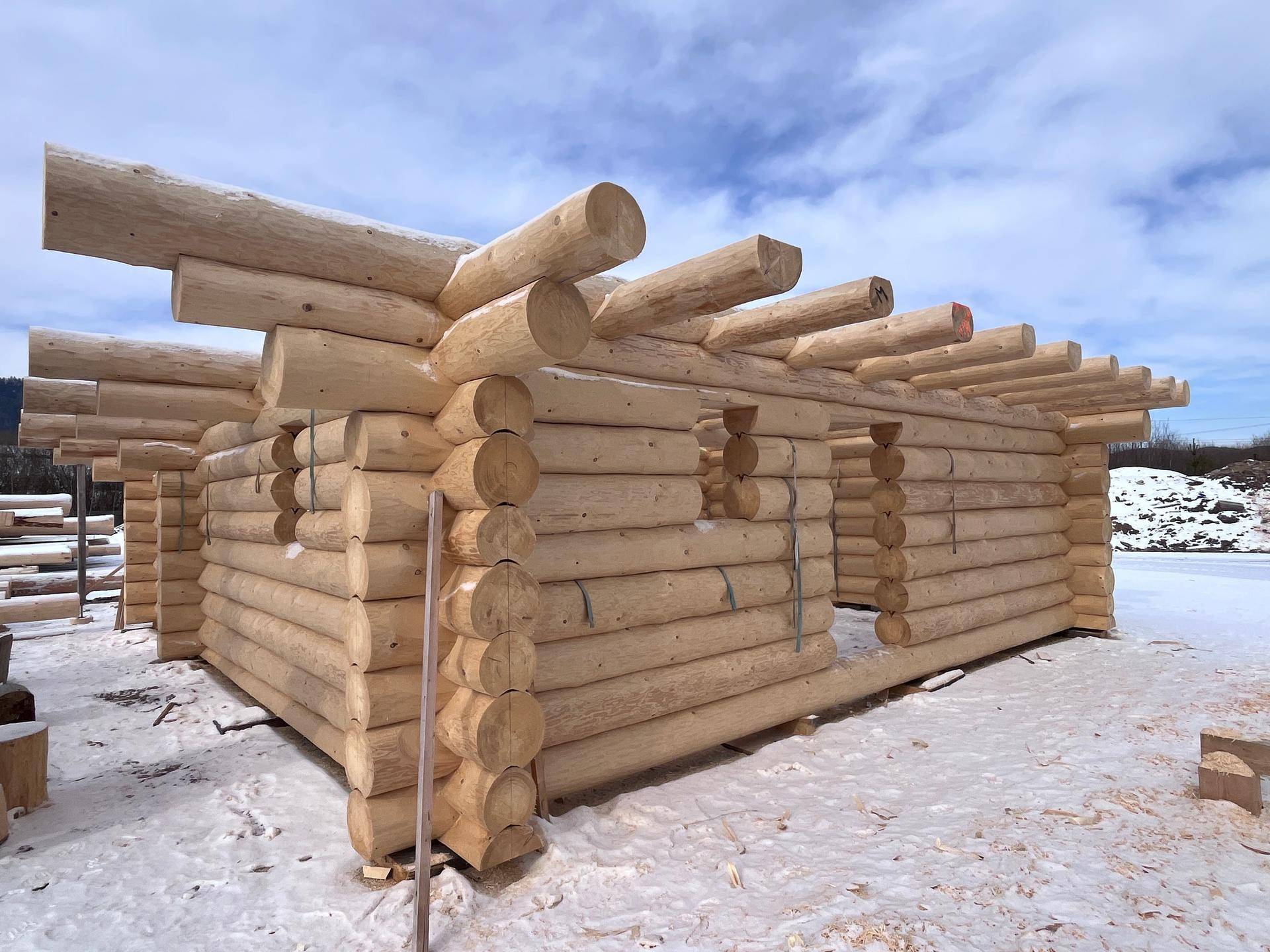 Baustelle eines Blockhauses im Winter mit frisch verlegten Holzstämmen und blauem Himmel.