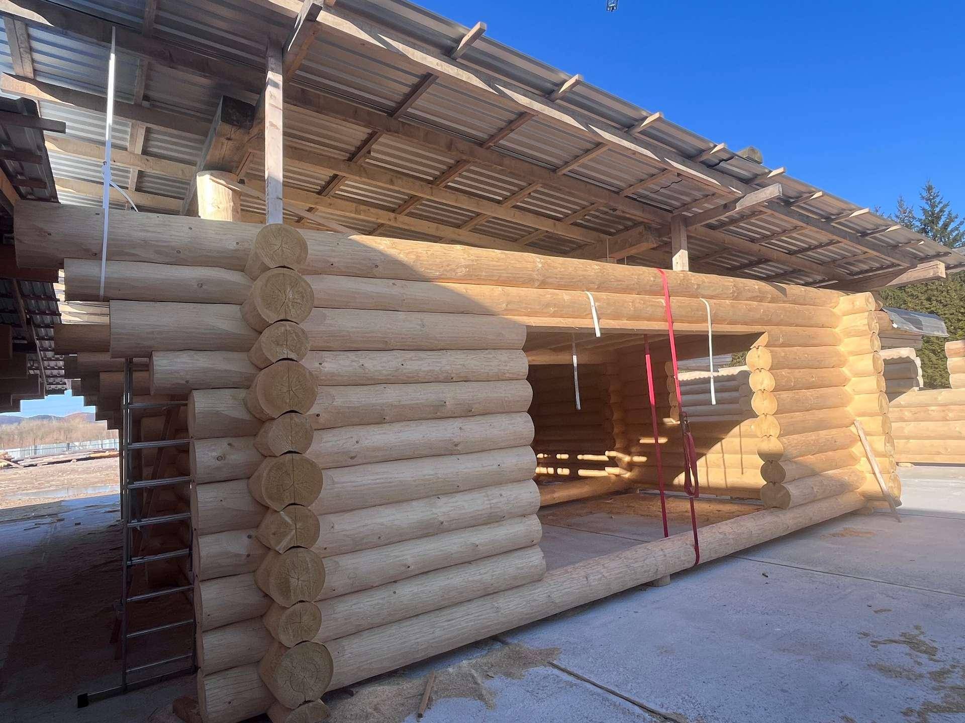 Blockhaus im Bau, mit sichtbaren Holzstämmen und provisorischem Dach, unter klarem Himmel.