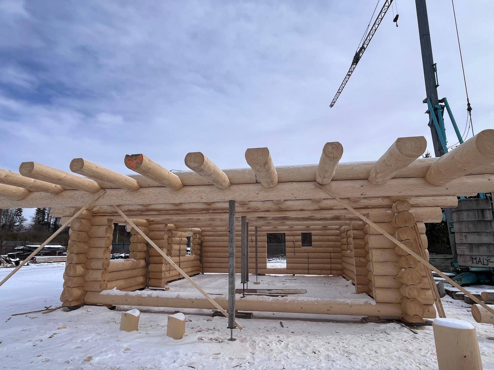 Rohbau eines Blockhauses im Winter mit sichtbaren Holzstämmen und Kran im Hintergrund.