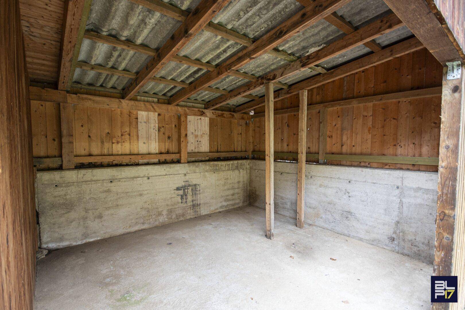 Einfacher Innenraum eines Holzschuppens mit Betonboden und sichtbaren Holzbalken.