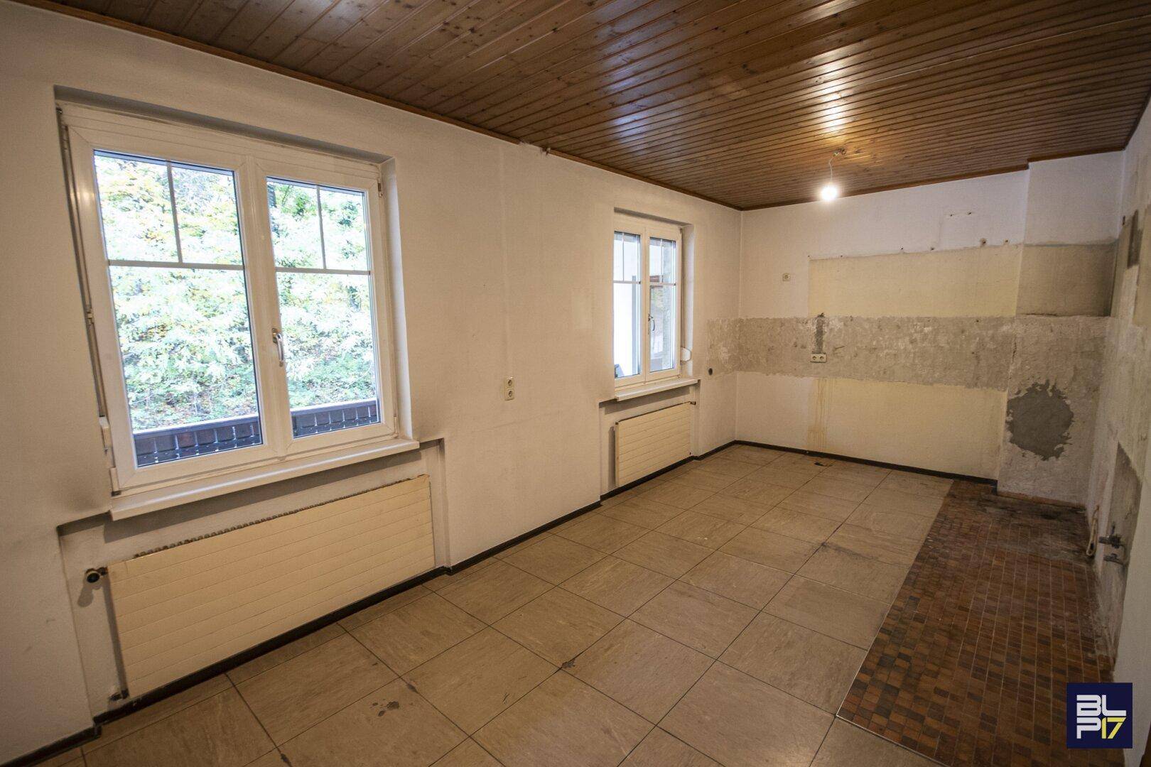Renovierungsbedürftige Küche mit Fliesenboden, Holzdecke und Blick ins Grüne.