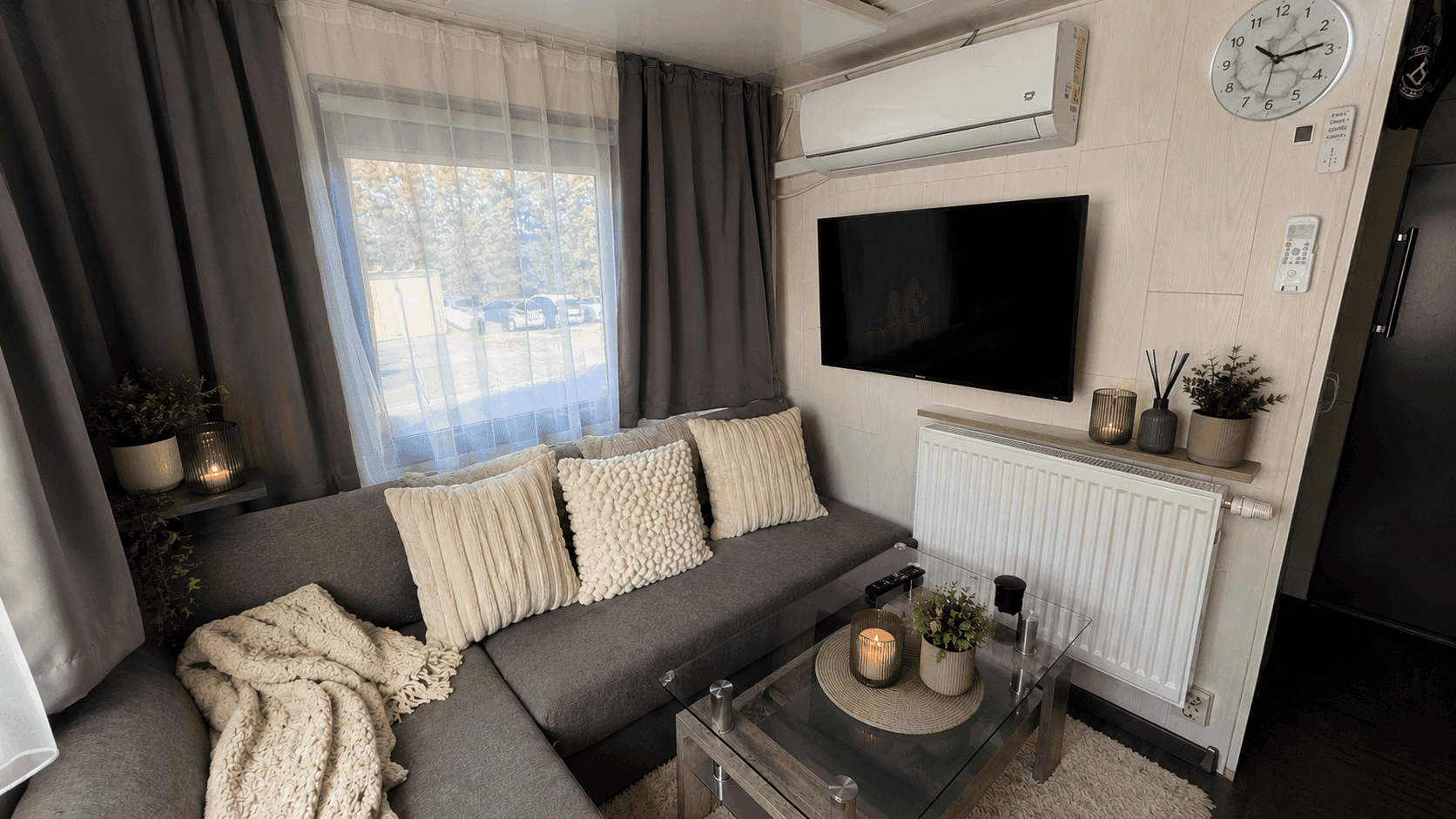Wohnzimmer mit grauem Ecksofa, Couchtisch, Wandfernseher, Heizkörper und Klimaanlage.