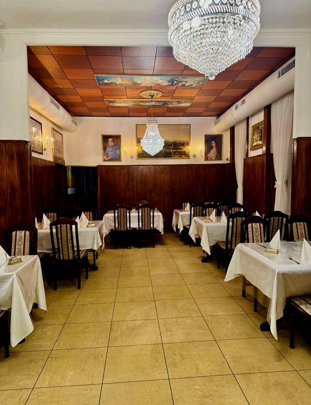 Großer Speisesaal mit mehreren Tischen, dunkler Holzvertäfelung und kunstvollen Deckenverzierungen, beleuchtet von einem prächtigen Kronleuchter.