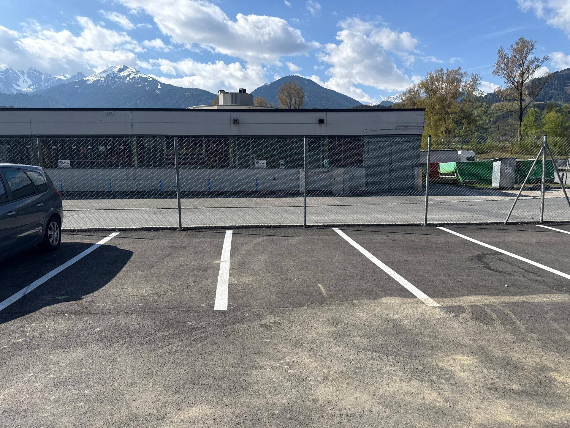 Geräumiger Parkplatz mit weißen Markierungen vor einem modernen Gebäude und beeindruckender Berglandschaft im Hintergrund.