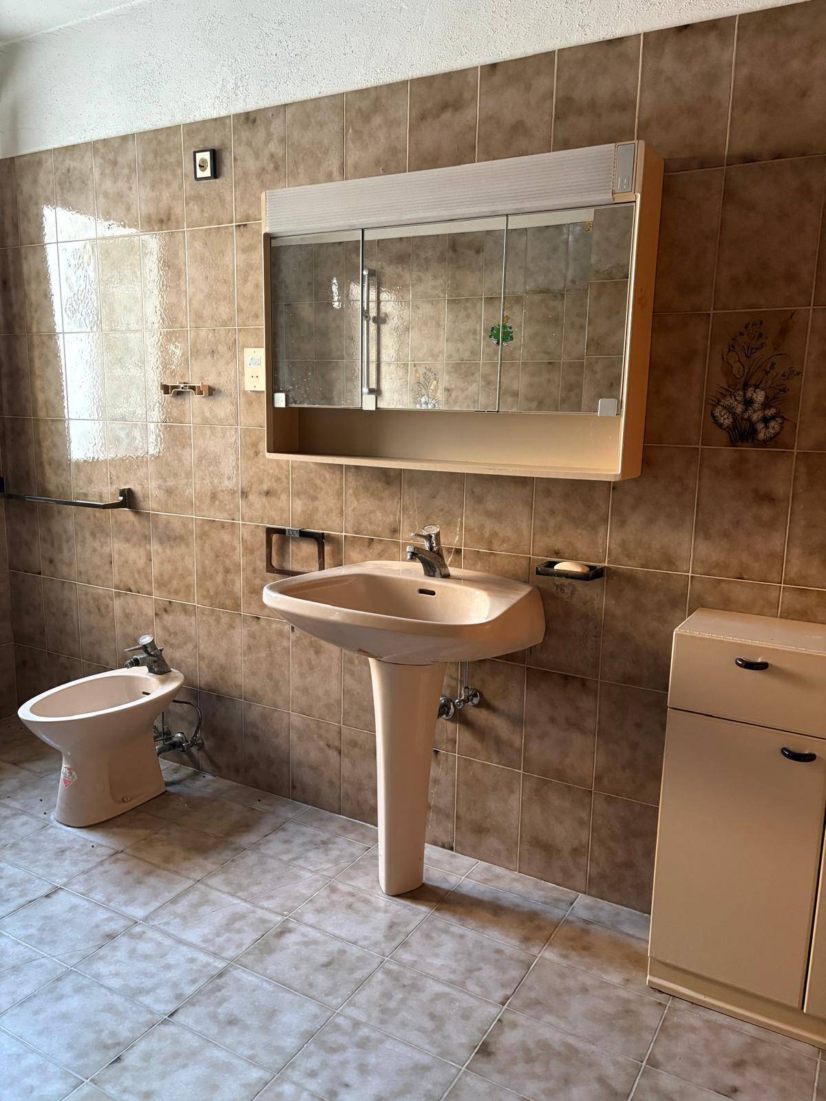 Badezimmer mit braunen Fliesen, Waschtisch, Bidet und Spiegelschrank.