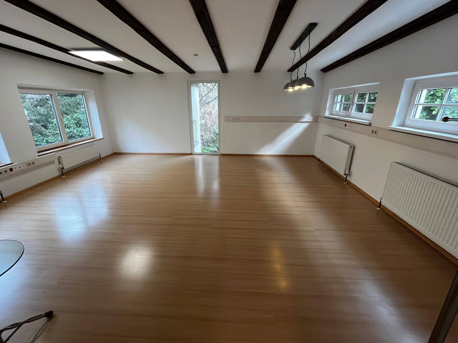 Heller Büroraum mit sichtbaren Holzbalken, Holzboden und Fenstern mit Blick ins Grüne.