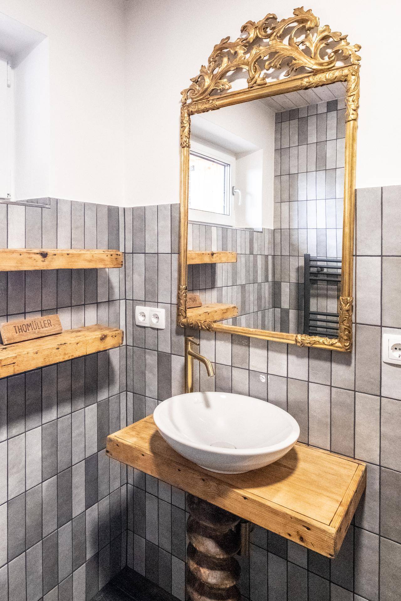 Modernes Badezimmer mit grauen Fliesen, Holzwaschtisch und goldenem Spiegel.