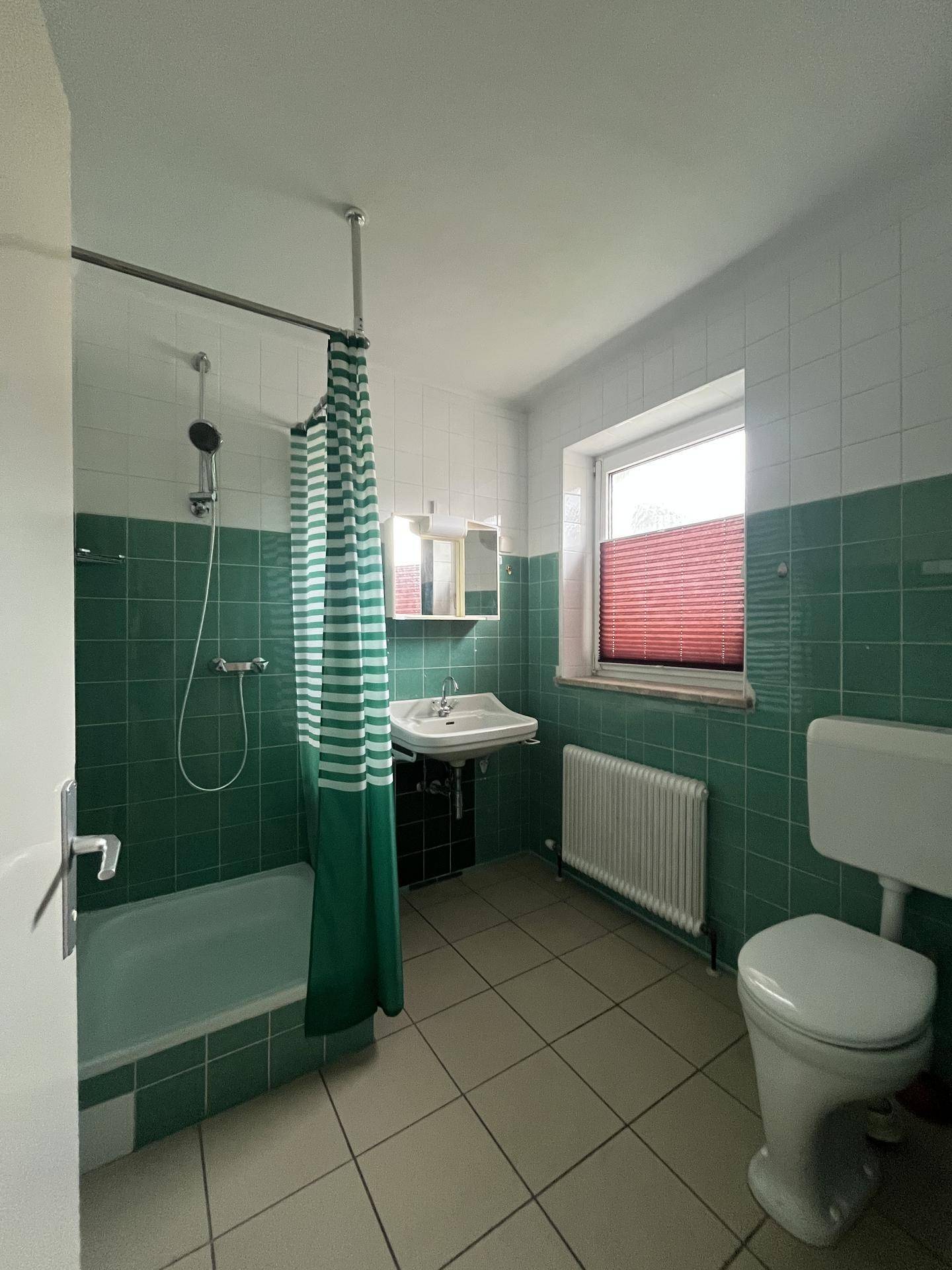 Badezimmer mit grünen Fliesen, Badewanne und Duschvorhang, sowie Fenster.