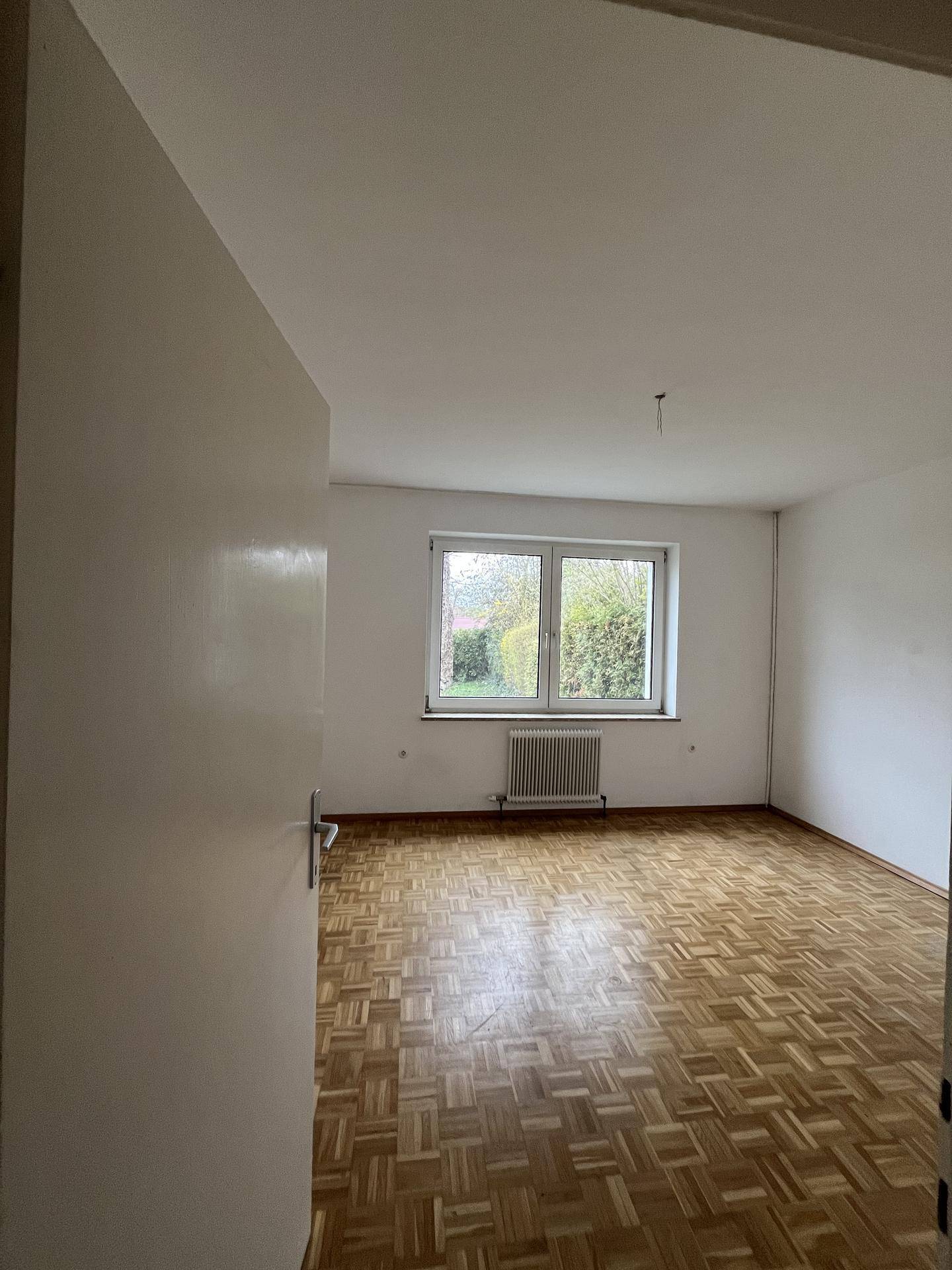 Leerer Raum mit klassischem Parkettboden und Fenster mit Blick ins Grüne.