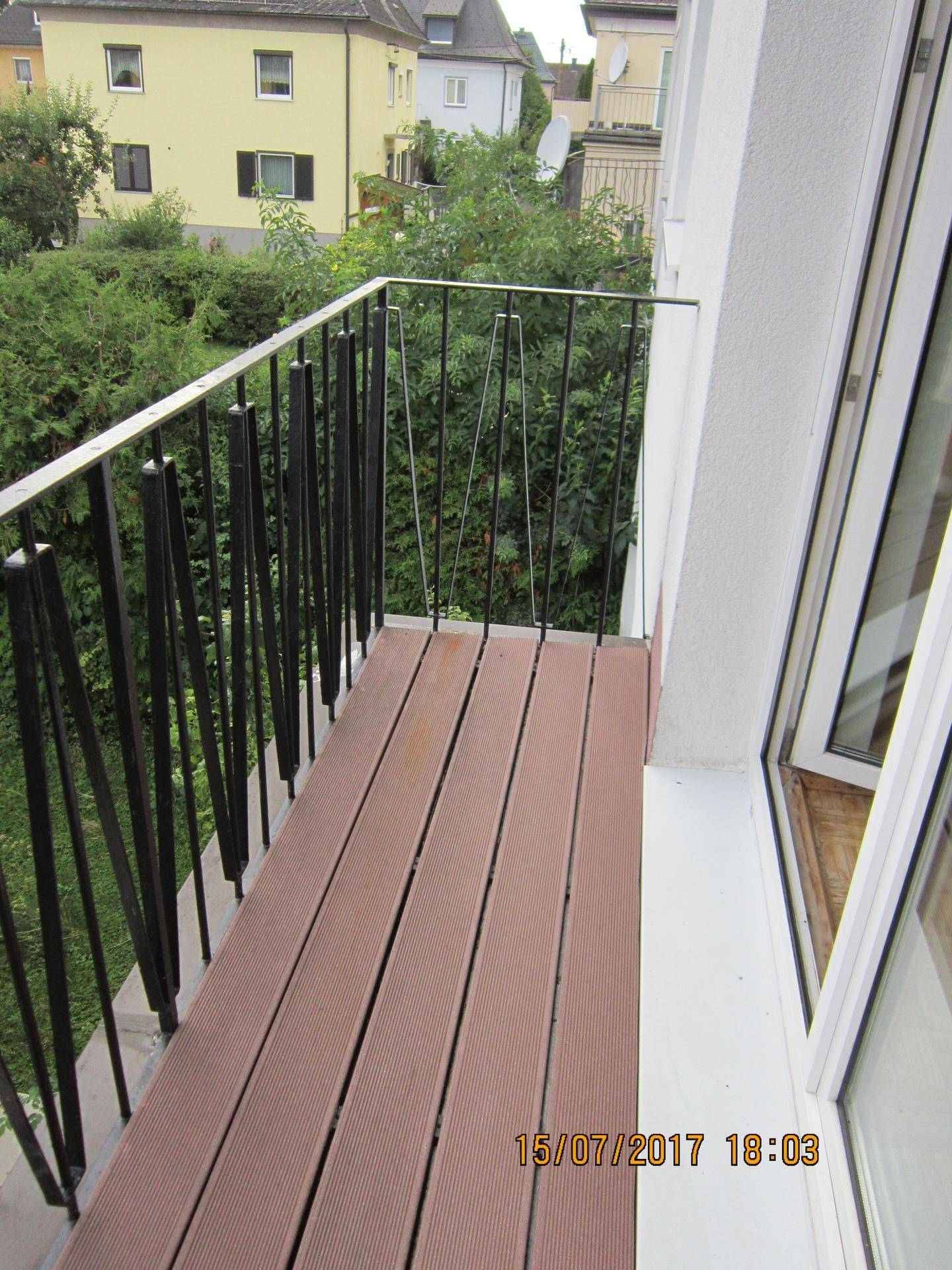 Kleiner Balkon mit Holzdielen und Metallgeländer, umgeben von grünen Hecken.