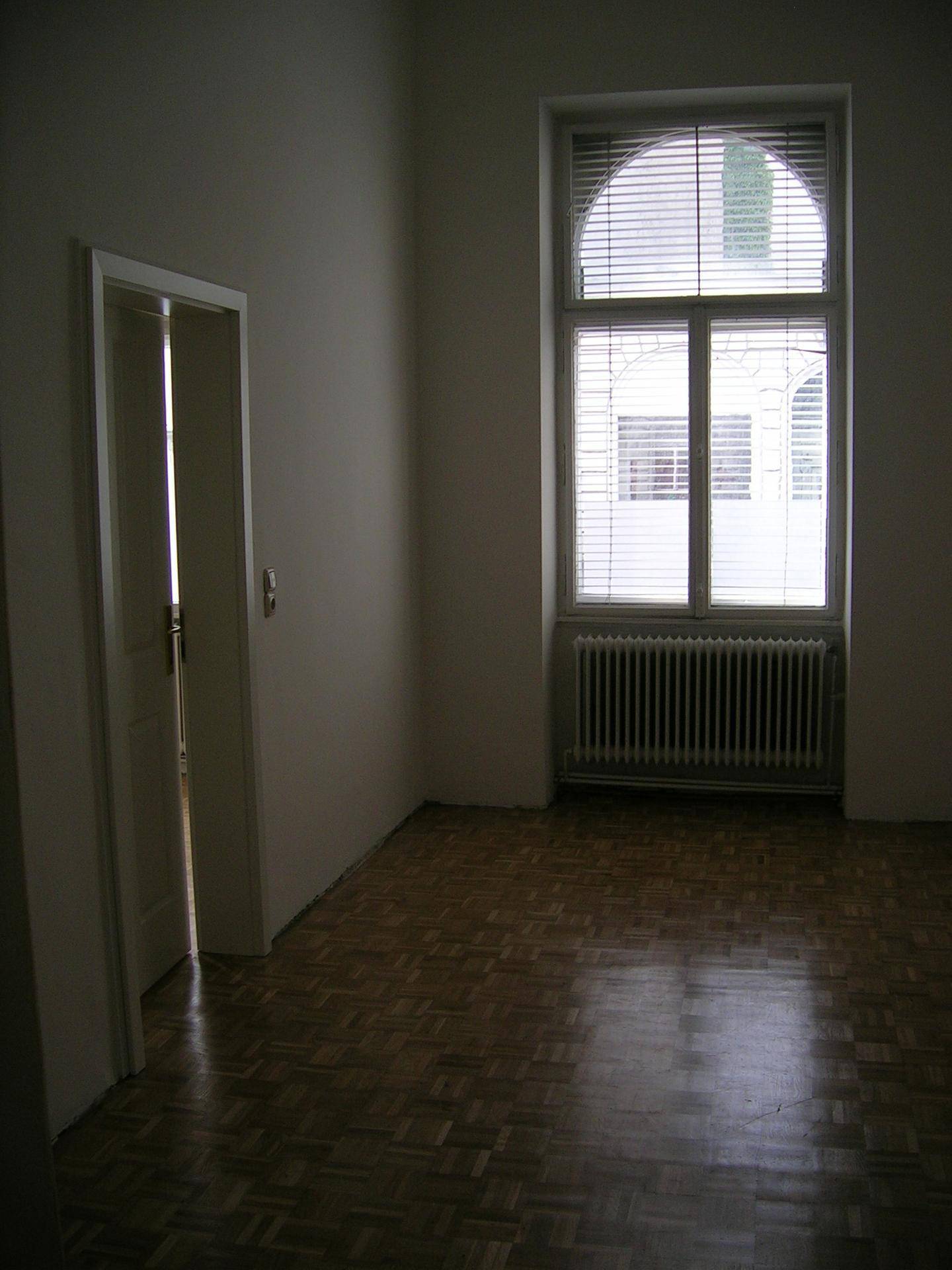 Leeres Zimmer mit Parkettboden, großem Fenster und Jalousien.