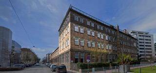 Großes mehrstöckiges Gebäude mit zahlreichen Fenstern in einer städtischen Umgebung mit Grünflächen.