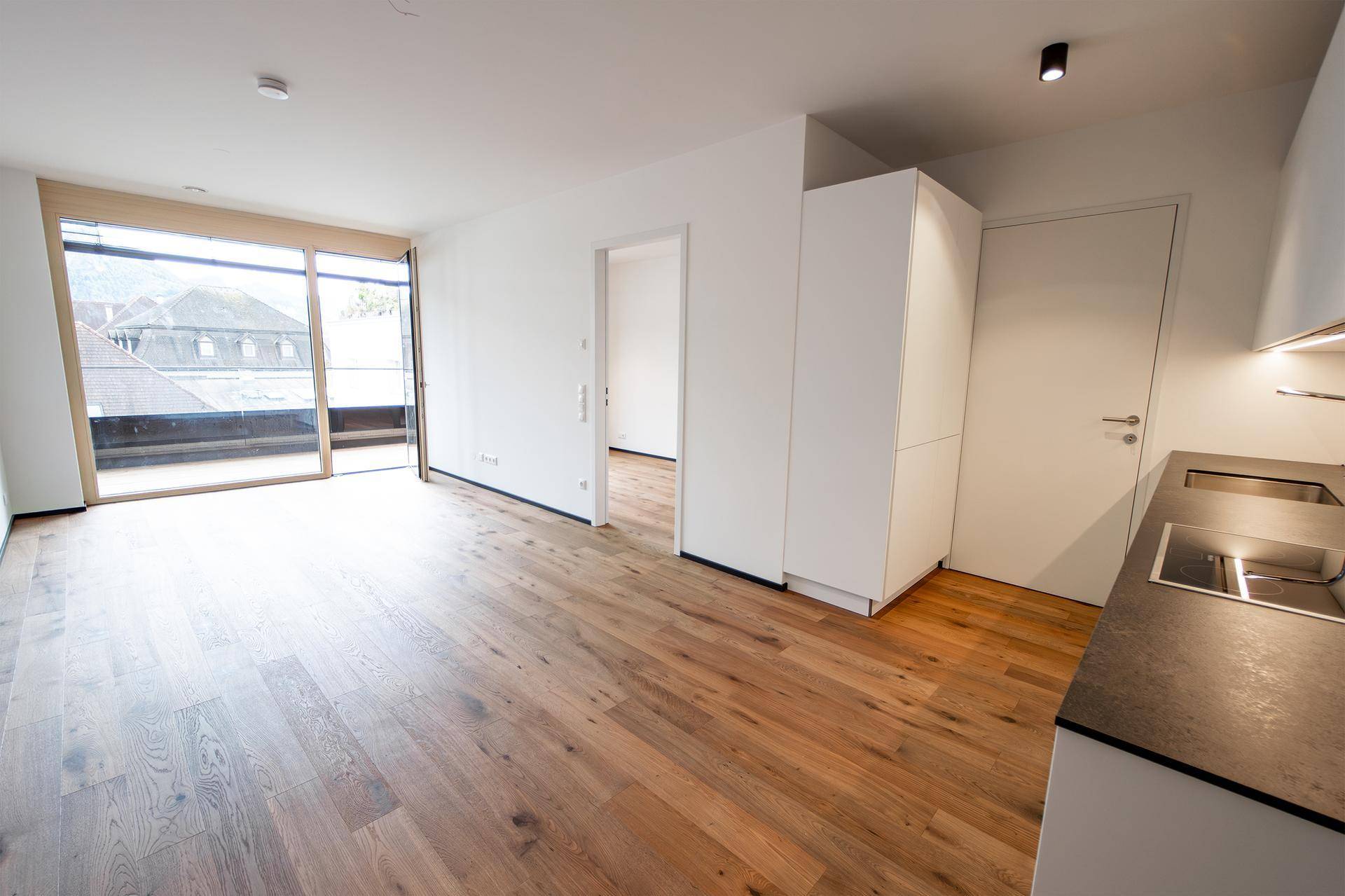 Geräumiger, heller Raum mit Holzboden und großem Fensterbereich, ideal für Wohn- und Essbereich.