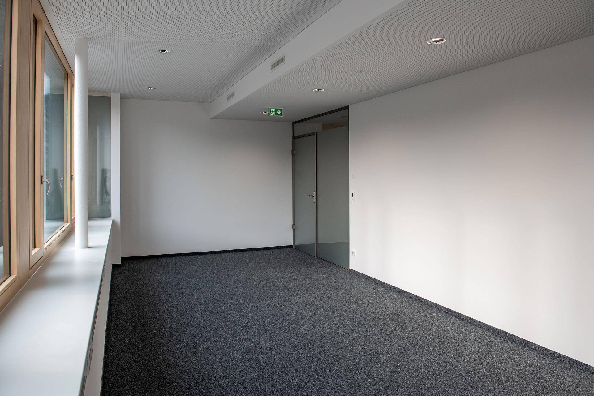 Moderner Büroraum mit grauem Teppichboden, weißen Wänden und einer Glastür zu einem angrenzenden Bereich.