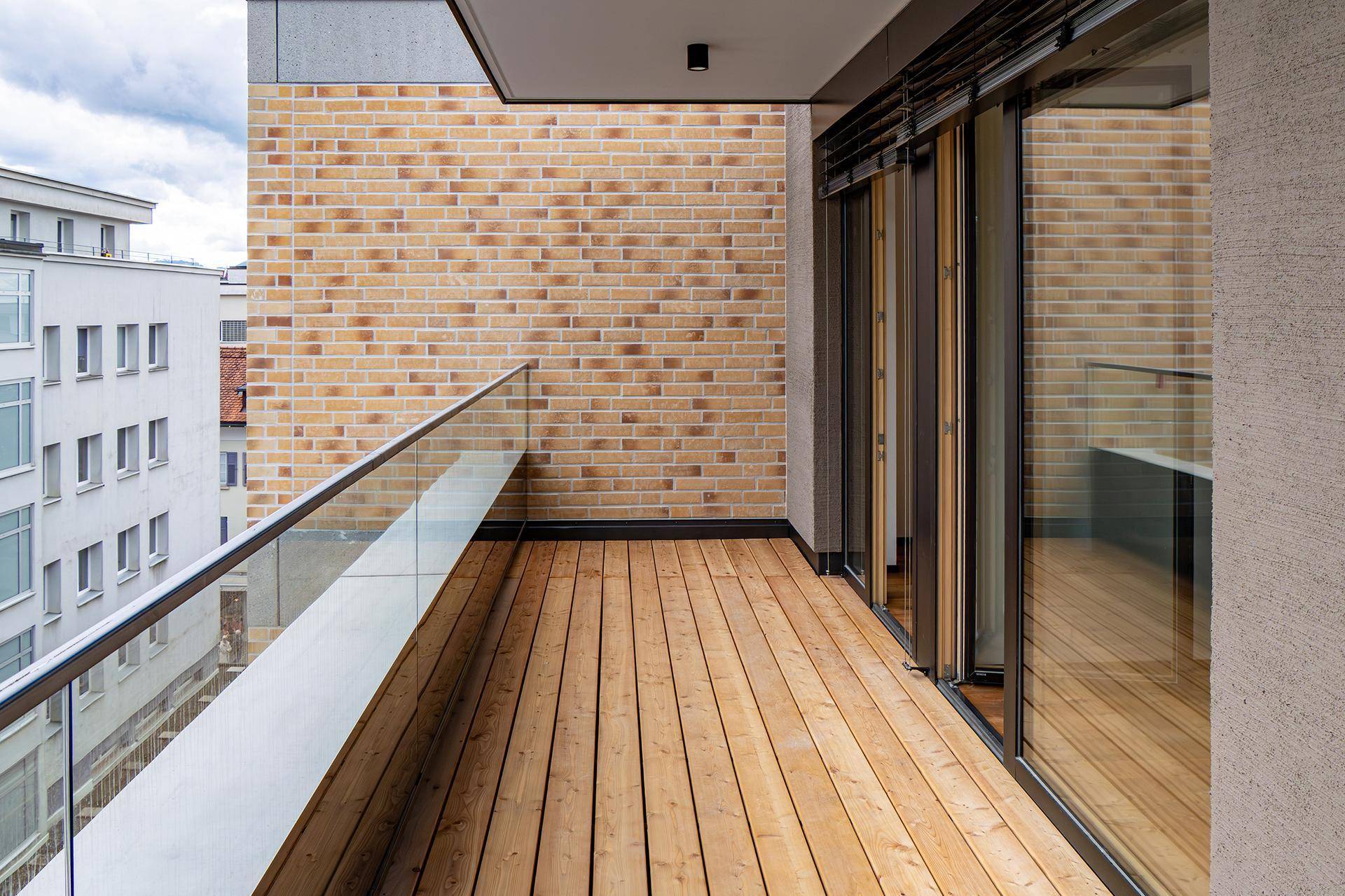 Geräumiger Balkon mit Holzfußboden, Glasgeländer und Blick auf die umliegenden Gebäude.