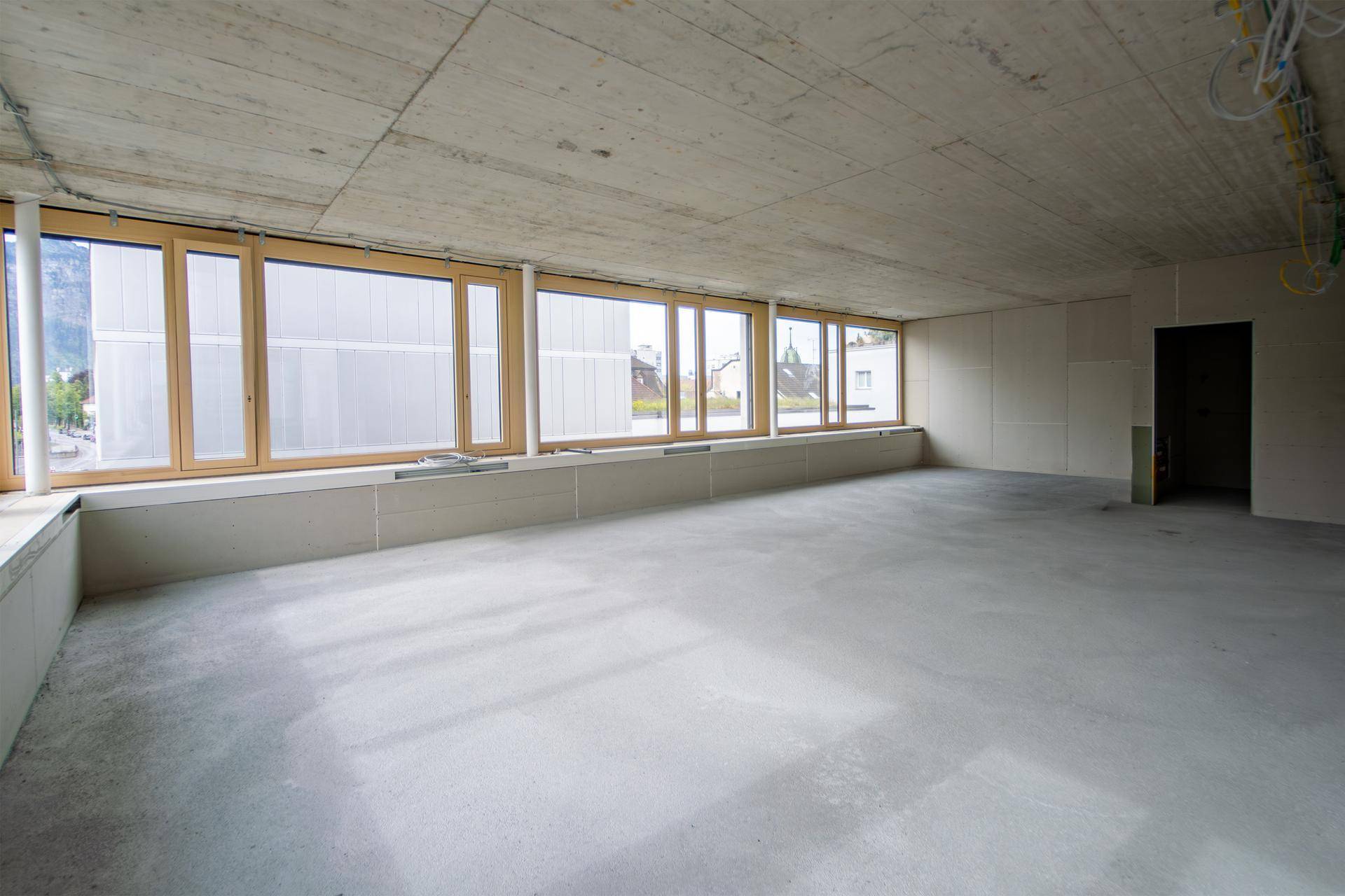 Unfertiger Innenraum mit Sichtbetonboden und -decke sowie breiten Holzfenstern.