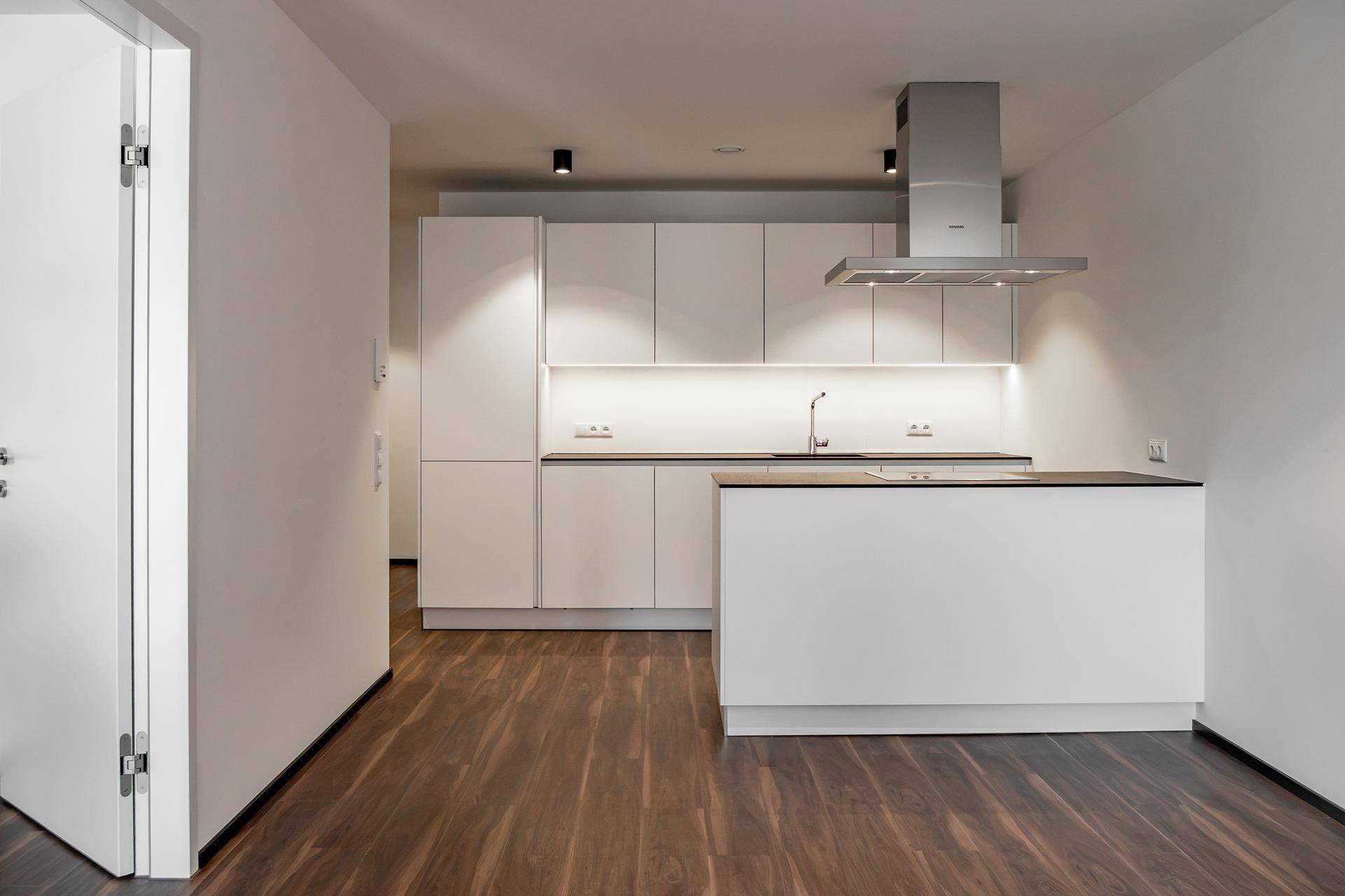 Moderne, unbenutzte Küche mit weißen Schränken, Dunstabzugshaube und dunkler Arbeitsplatte.