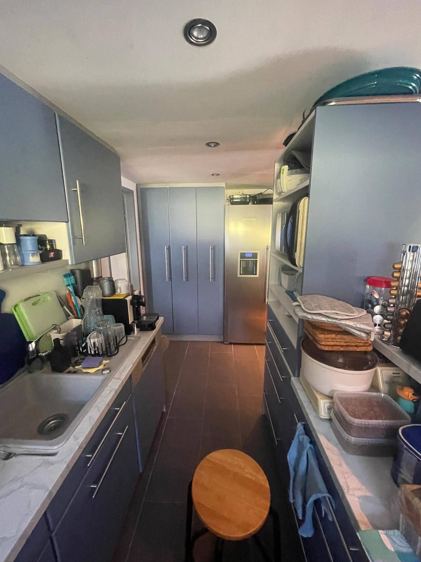 Schmale Küche mit blauen Schränken, viel Stauraum und einem großen Kühlschrank am Ende.