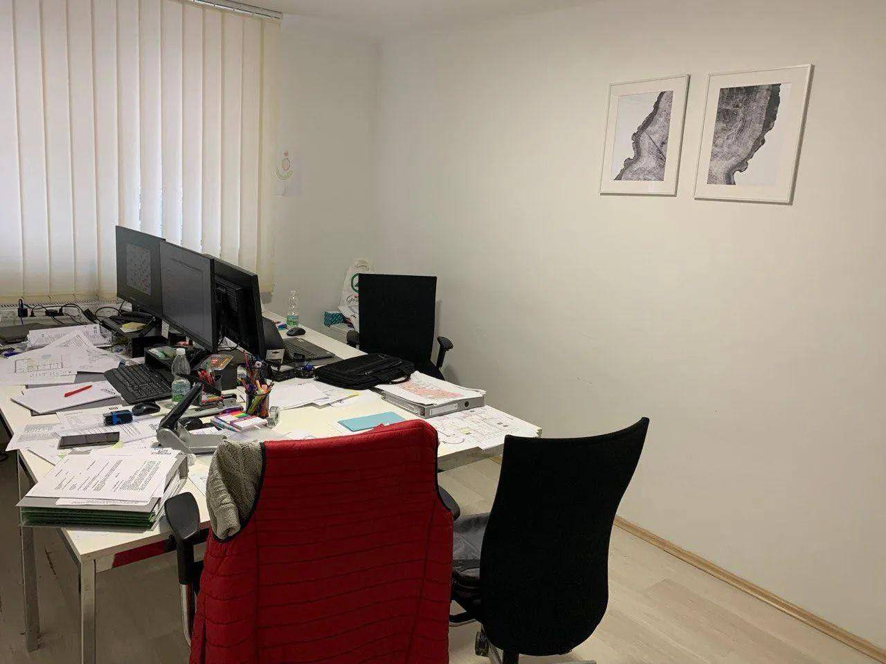 Arbeitsbereich mit mehreren Computern, Schreibtischen und Dokumenten, beleuchtet durch ein großes Fenster.