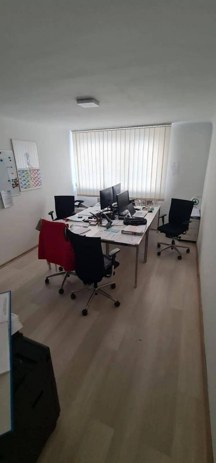 Geräumiges Büro mit mehreren Arbeitsplätzen, Computern und einem großen Fenster mit Jalousien.