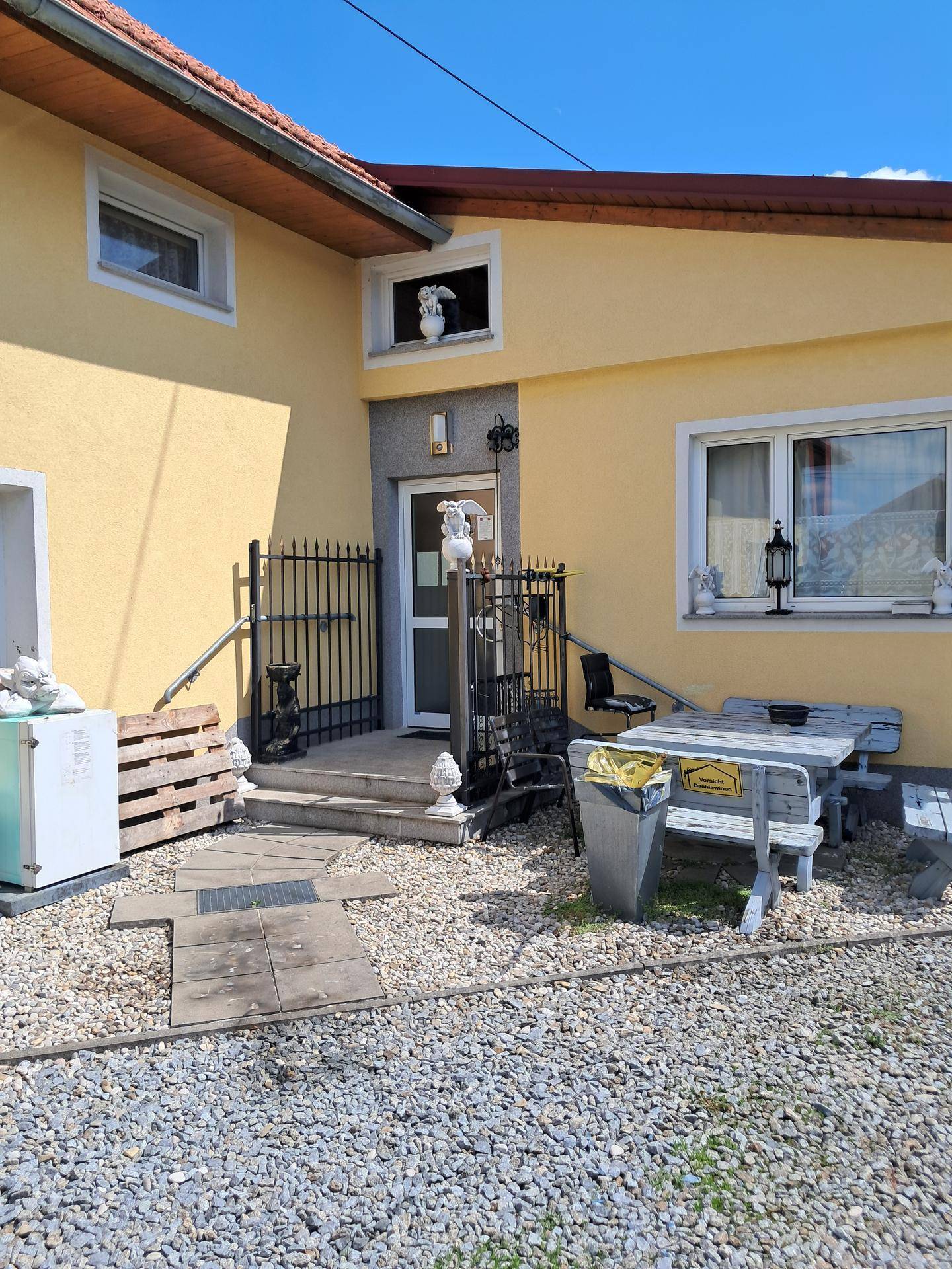 Eingangsbereich des Hauses mit gelber Fassade, Schotterfläche und einem einfachen Picknicktisch.