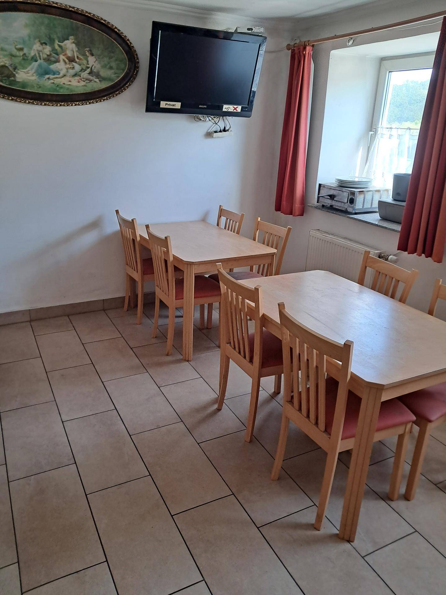 Esszimmer mit mehreren Holztischen und Stühlen, Fliesenboden und einem Wandfernseher.