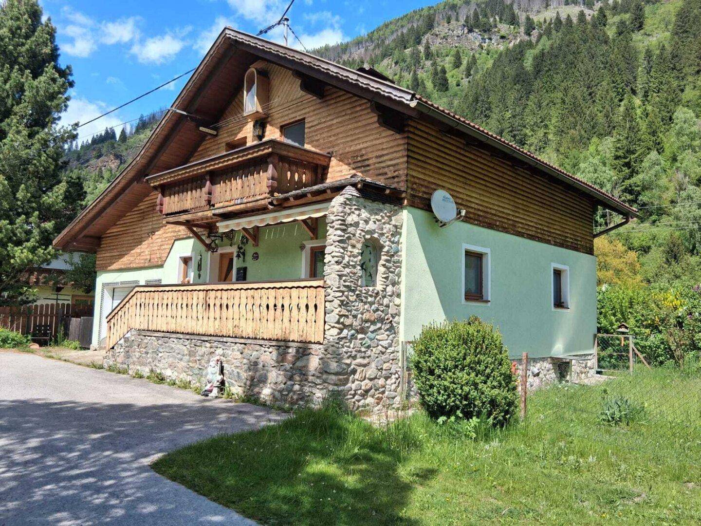 Alpines Haus mit Holzverkleidung, Steinsockel und Balkon vor bergiger Kulisse.