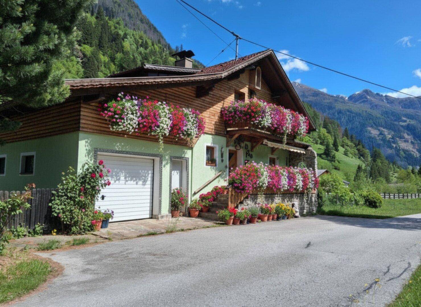 Traditionelles Haus mit blühenden Blumenkästen an Balkonen und Fenstern in alpiner Umgebung.