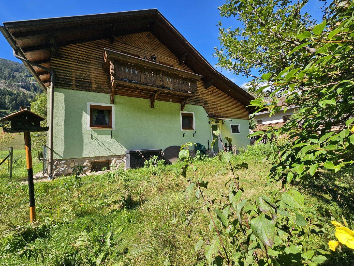 Haus mit Balkon, umgeben von einem naturbelassenen Garten und Bergpanorama.