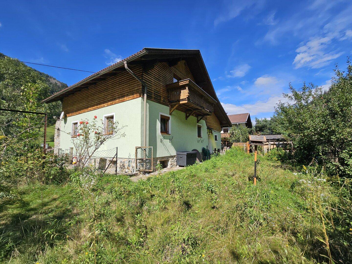 Haus mit Balkon und Garten, umgeben von üppiger Vegetation und Bergen.