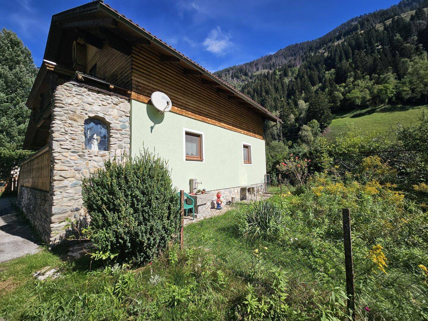 Seitenansicht des Hauses mit Steinsockel und Garten, umgeben von Natur.